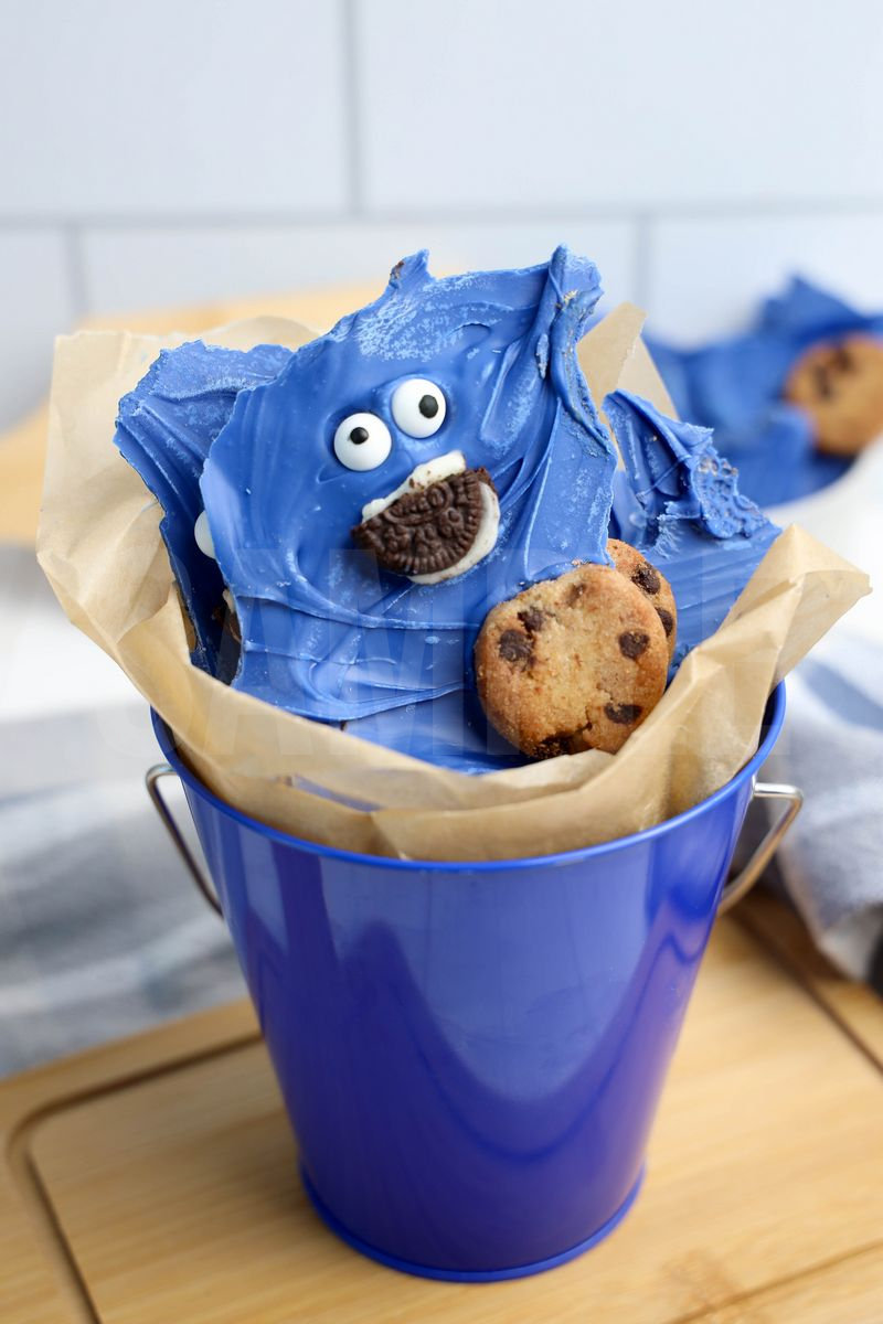 The Cookie Monster Bark comes in a blue pail with parchment paper with a blue and white striped cloth on a white wood backdrop.