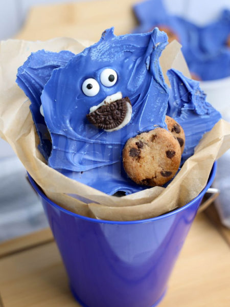 The Cookie Monster Bark comes in a blue pail with parchment paper with a blue and white striped cloth on a white wood backdrop.