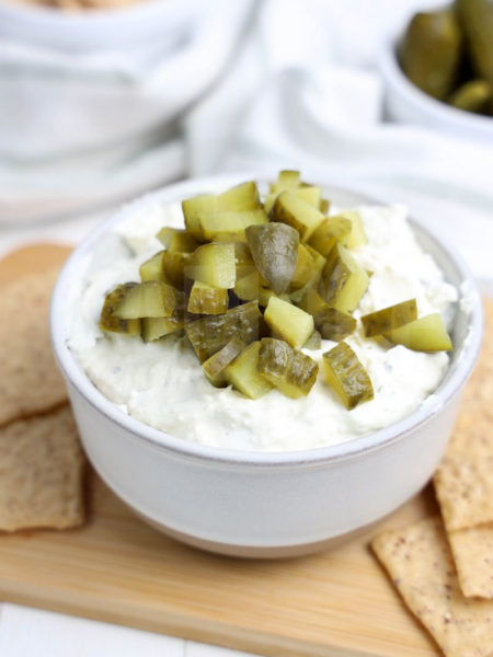 The Dill Pickle Dip comes in a cream bowl on a wood cutting board with a light green and white striped cloth on a white wood backdrop.