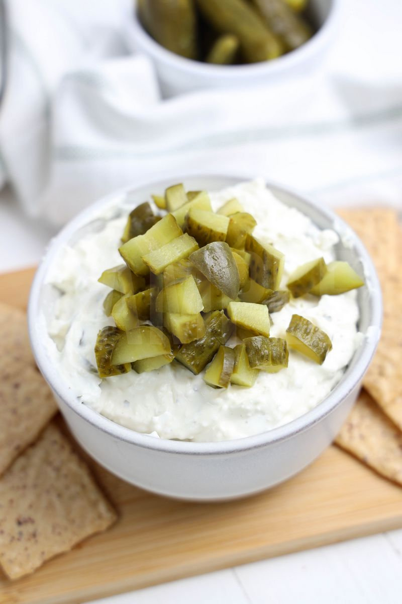 The Dill Pickle Dip comes in a cream bowl on a wood cutting board with a light green and white striped cloth on a white wood backdrop.