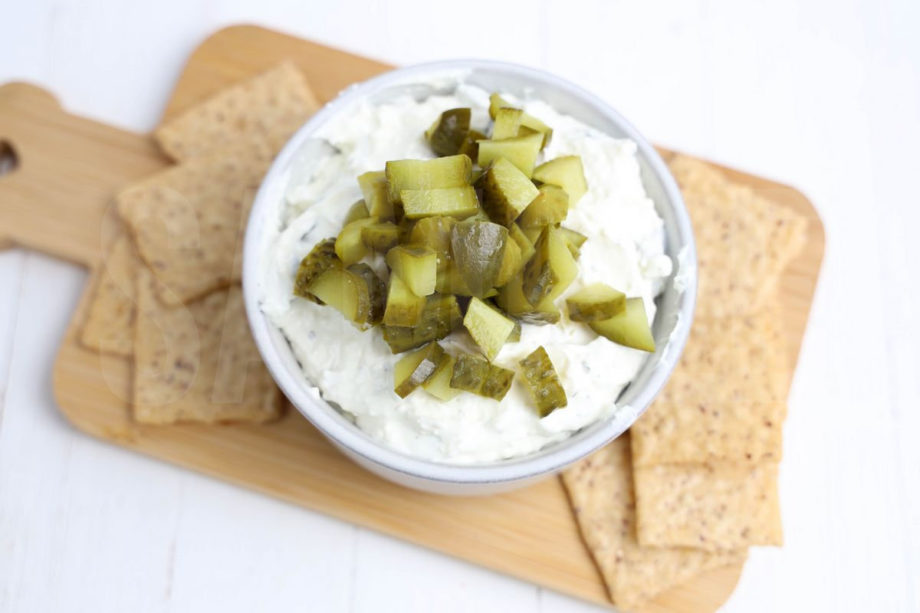 The Dill Pickle Dip comes in a cream bowl on a wood cutting board with a light green and white striped cloth on a white wood backdrop.