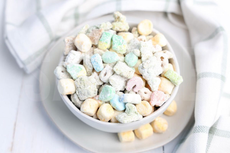 The Lucky Charms Puppy Chow comes in a white bowl on a white plate with a light green and white plaid cloth on a white wood backdrop.