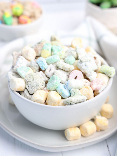 The Lucky Charms Puppy Chow comes in a white bowl on a white plate with a light green and white plaid cloth on a white wood backdrop.