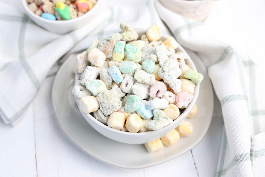 The Lucky Charms Puppy Chow comes in a white bowl on a white plate with a light green and white plaid cloth on a white wood backdrop.