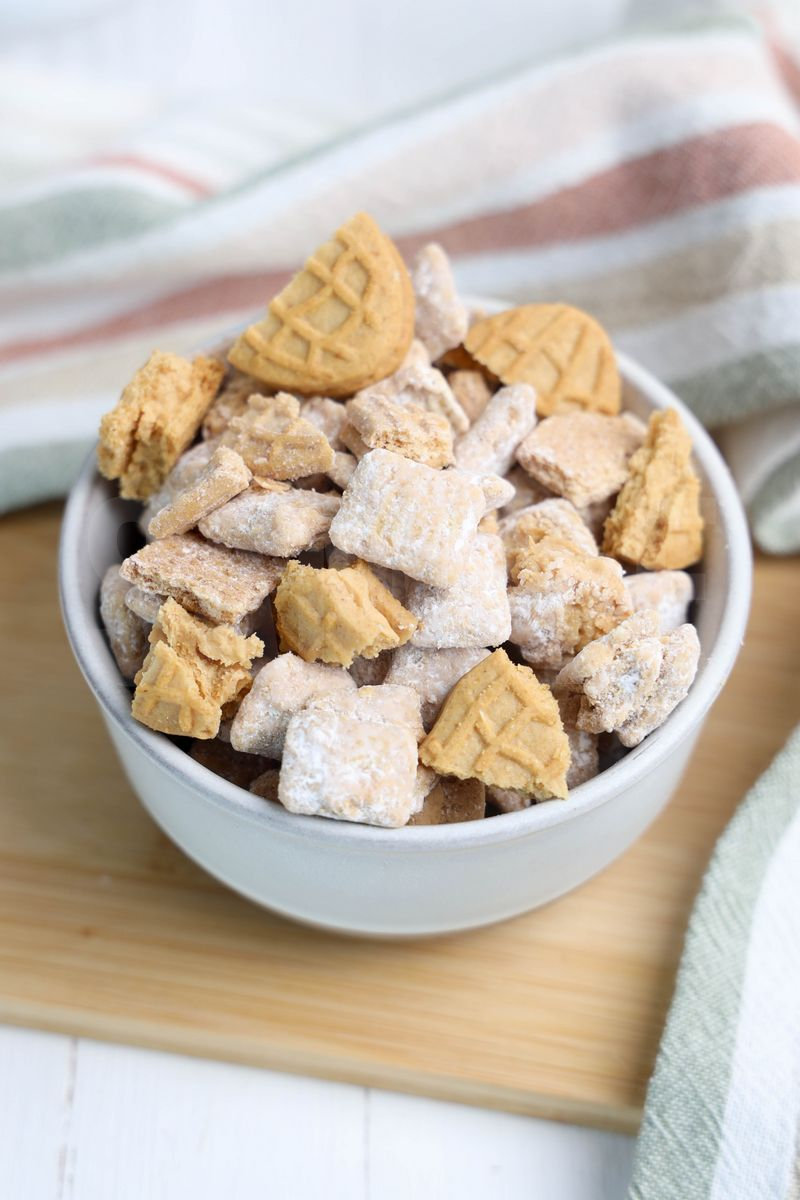 The Nutter Butter Puppy Chow comes in a cream stone bowl with wood cutting board and rainbow cloth on white wood backdrop.