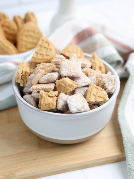 The Nutter Butter Puppy Chow comes in a cream stone bowl with wood cutting board and rainbow cloth on white wood backdrop.