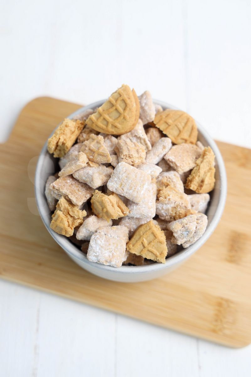 The Nutter Butter Puppy Chow comes in a cream stone bowl with wood cutting board and rainbow cloth on white wood backdrop.