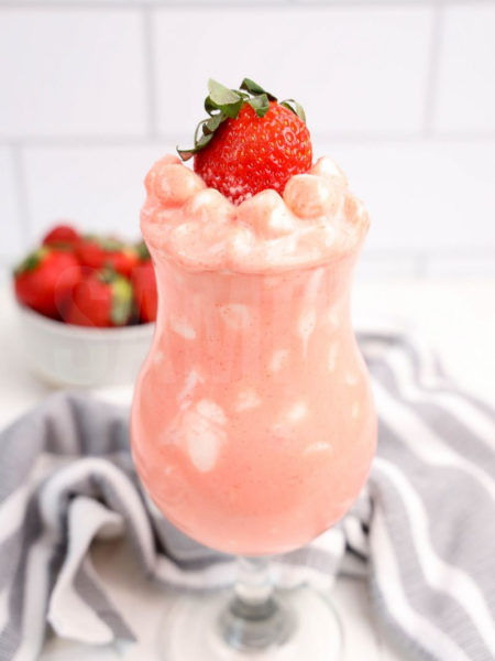 The Strawberry Fluff comes in a clear glass with a thick gray and white striped cloth on a white wood backdrop.