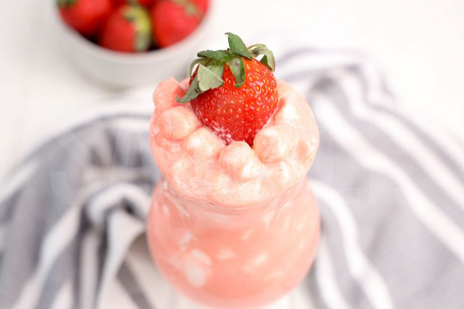The Strawberry Fluff comes in a clear glass with a thick gray and white striped cloth on a white wood backdrop.