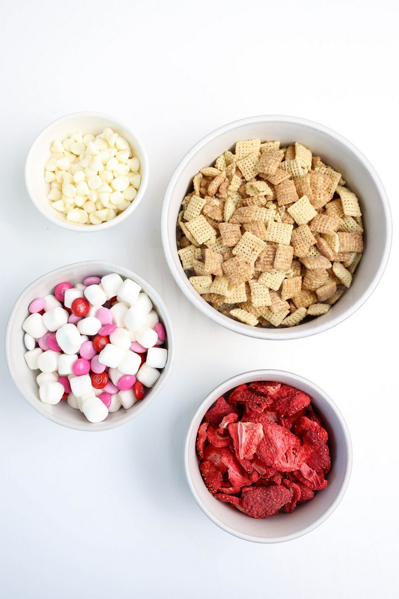 The Strawberry Puppy Chow comes in a white bowl on a white plate with a light gray and white striped cloth on a white wood backdrop.