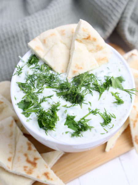 The Tzatziki Dip comes in a white bowl on a wood cutting board with a olive green cloth on a white wood backdrop.