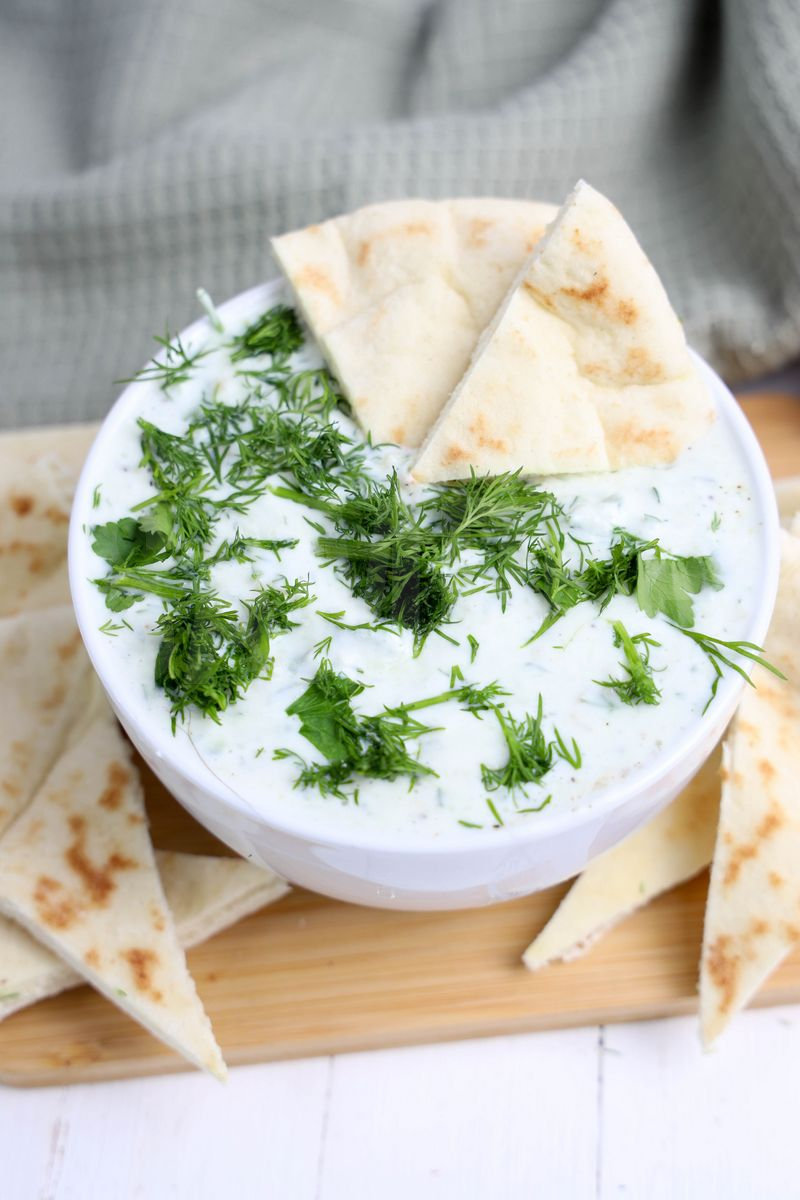 The Tzatziki Dip comes in a white bowl on a wood cutting board with a olive green cloth on a white wood backdrop.