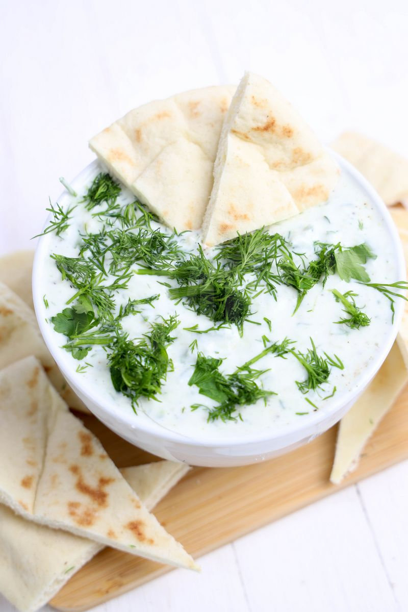 The Tzatziki Dip comes in a white bowl on a wood cutting board with a olive green cloth on a white wood backdrop.