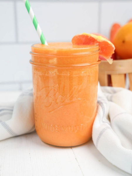 The Carrot Apple Orange Smoothie comes in a mason jar with a grey and white plaid cloth on white wood backdrop.