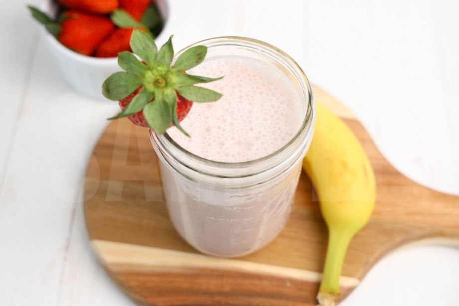 The Strawberry Banana Smoothie comes in a mason jar with a grey and white plaid cloth on white wood backdrop.