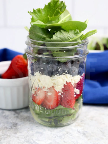 The July 4th Mason Jar Salad comes in a clear glass mason jar with marble wood backdrop.